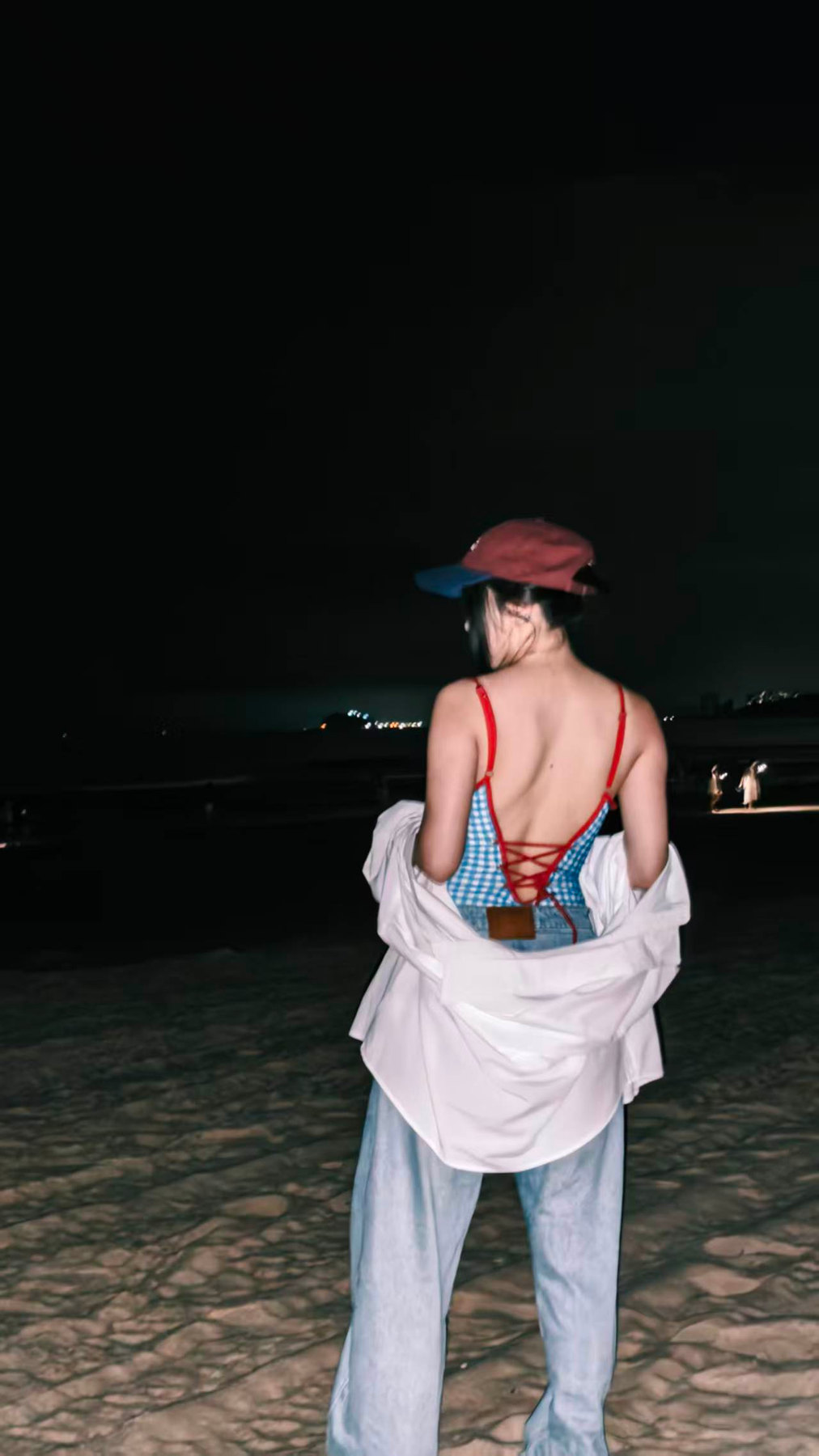 夜里的沙滩上,戴红色帽子的女孩背对镜头,白衬衫滑到手肘